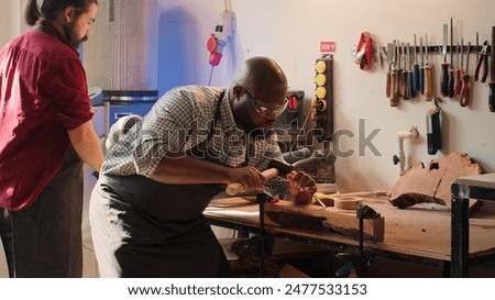 Sculptor shaping raw timber using chisel and hammer in carpentry shop with coworker cleaning workbench in background. BIPOC man making wood sculptures, engraving plank with tools, camera A