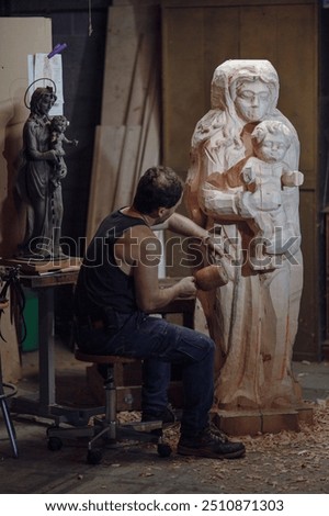 Sculptor is using a mallet and chisel to carve a large wooden statue of the virgin mary with child in his workshop
