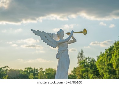 sculpture of angel blowing golden horn on sunrise sky clouds with trees.
A trumpeting golden music angel statue detail.