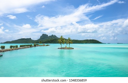 Sculpture Of Human Sitting In The Middle Of The Isolated Island With Two Palm Trees In Beautiful Lagoon And The Big Island On The Background. Airport Bora Bora, South Pacific, Tahiti French Polynesia