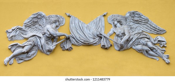 Sculptures, Stucco In The Style Of Classicism, Bas-relief. Two Angels Crossed Flags On A Yellow Background. Gate Protection From Unwanted Guests
