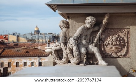 Sculptures of two kids with wreaths and scepters in their hands. Next to it is a bas-relief of a vase with fruit. Decoration of building. Saint Isaacs Cathedral on background. St Petersburg, Russia.