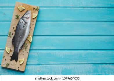 Sea Bass On Kitchen Table With Lemon And Rosemary On Blue Wooden Background. Top View With Space To Write.