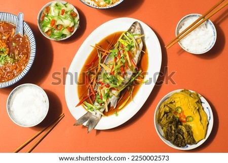  Sea bass or Spigola fish steamed with ginger, red peppers in soy sauce served  with Chinese cabbage, traditional spicy soup and rice. Chinese New Year, festive table in Cantonese style. Red terracott