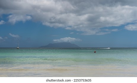 Sea idyll on a sunny day. A yacht is visible in the calm turquoise ocean. The outline of the island on the horizon. Clouds in the blue sky. Seychelles. Mahe. Beau Vallon - Shutterstock ID 2253753635