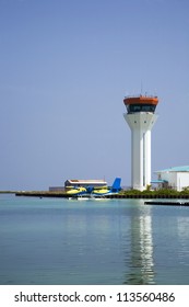 A Sea Plane Lands On The Water With The Air Traffic Control Tower In The Background.