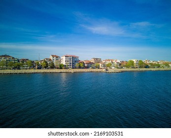 Sea Promenade With People Walking Near Deep Calm Cool Black Sea, Against Backdrop Of Ancient Resort Small Town Of Pomorie In Bulgaria Under A Cloudy Soft Blue Sky