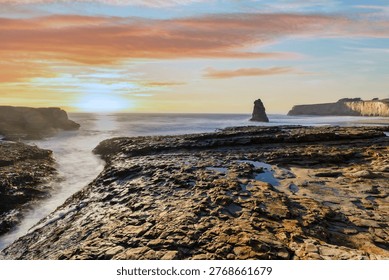 A sea stack rises from the Pacific Ocean at Davenport Beach, California, as soft waves and warm sunset light reflect on wet sand, creating a calm and scenic coastal view