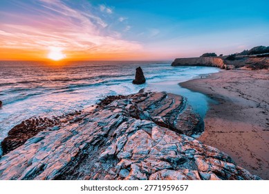 A sea stack rises from the Pacific Ocean at Davenport Beach, California, as soft waves and warm sunset light reflect on wet sand, creating a calm and scenic coastal view