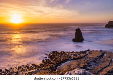 A sea stack rises from the Pacific Ocean at Davenport Beach, California, as soft waves and warm sunset light reflect on wet sand, creating a calm and scenic coastal view