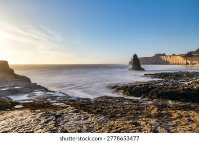 A sea stack rises from the Pacific Ocean at Davenport Beach, California, as soft waves and warm sunset light reflect on wet sand, creating a calm and scenic coastal view
