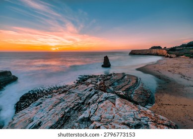 A sea stack rises from the Pacific Ocean at Davenport Beach, California, as soft waves and warm sunset light reflect on wet sand, creating a calm and scenic coastal view
