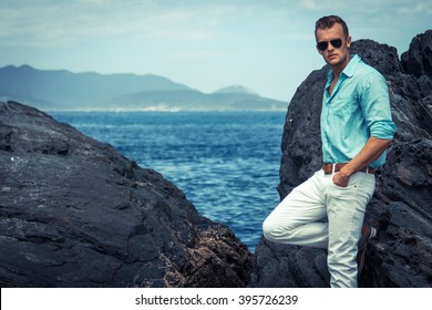 Sea/beach Fashion Concept. Portrait Of A Young And Handsome Man In A Aqua Shirt, White Pants And Trendy Glasses Posing Over The Shore Rocks Of A White Sand Beach. Close Up. 