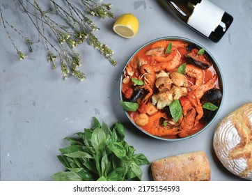 Seafood cacciucco is a popular dish in Italy. Thick tomato soup with shrimps and mussels in a round plate. Ready meal, ciabatta, bottle of wine, herbs, lemon and herbs on a gray table - Shutterstock ID 2175154593