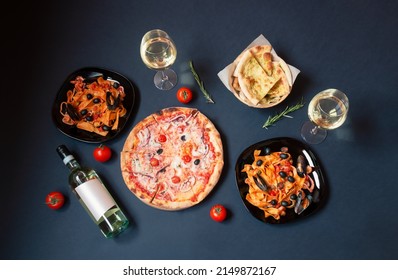 Seafood pizza, plates of pappardelle pasta with seafood with tomato sauce, bottle of white wine, glasses of white wine and focaccia bread on dark background. Top view. - Powered by Shutterstock