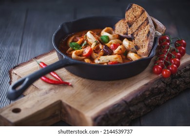 Seafood sate with squid, beans and tomatoes in a black pan on a black wooden background with toasted bread