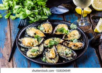 Seafood. Shellfish mussels. Baked mussels with cheese, cilantro and lemon in shells on cast iron black plate. Plate of mussels, cold white wine, lemon and cilantro on old wooden blue background.