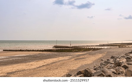 A seafront with breakwaters stretching out towards the sea. The sandy beach is covered in shingle and large rocks are in the foreground.