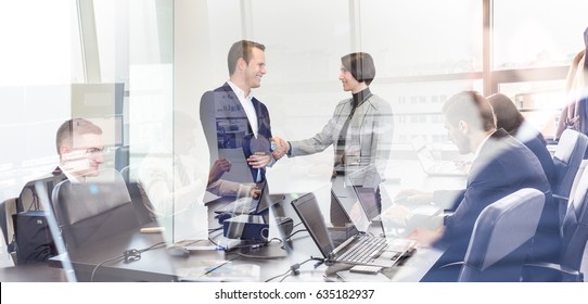 Sealing A Deal. Business People Shaking Hands, Finishing Up Meeting In Corporate Office. Businessmen Working On Laptop Seen In Glass Reflection. Business And Entrepreneurship Concept.