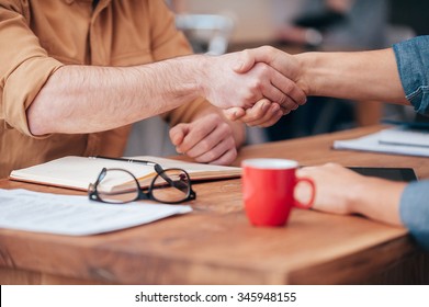 Sealing a deal. Close-up of two men shaking hands while sitting at the wooden desk 