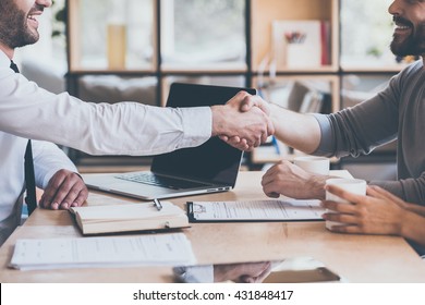 Sealing a deal. Side view close-up of two young man shaking hands while sitting at the wooden desk