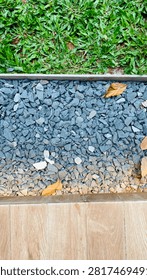 Seamlessly blending natural elements with modern design, the outdoor pathway features rich wooden-patterned ceramic tiles flanked by a bed of smooth, dark river pebbles and lush green grass. The textured warmth of the wood-look ceramics provides a clean, sophisticated surface to walk on, while the coarse pebbles introduce an organic, grounding contrast in color and tactile feel. Bright blades of grass fringe the edges, softening the stone borders and bringing a vibrant burst of life to the composition. Together, these three materials harmonize into a stylish, serene landscape that feels both thoughtfully curated and effortlessly rooted in nature.