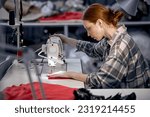 seamstress lady in casual wear sit behind table using sewing machine, making modern clothes, young redhead caucasian woman at workshop, holding red fabric, enjoy tailoring, controlling equipment