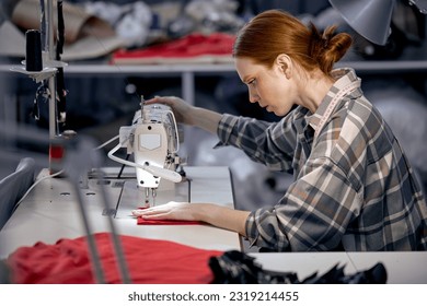 seamstress lady in casual wear sit behind table using sewing machine, making modern clothes, young redhead caucasian woman at workshop, holding red fabric, enjoy tailoring, controlling equipment