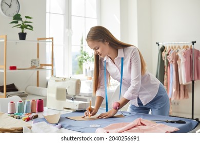Seamstress at work. Dressmaker making clothes in modern studio. Tailor holding pencil and marking fabric. Woman standing at table with cut textile, sewing machine, thread, pins, needles, tape, cutouts