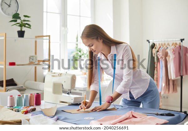 Seamstress at work. Dressmaker making clothes in\
modern studio. Tailor holding pencil and marking fabric. Woman\
standing at table with cut textile, sewing machine, thread, pins,\
needles, tape,\
cutouts