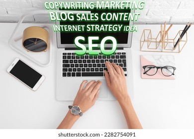 Search engine optimization (SEO). Copywriter working on laptop at white table, top view. Many words coming out of computer