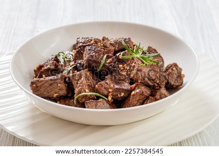 seared juicy new york strip steak bites with butter, rosemary and garlic in bowl on white wood table, close-up
