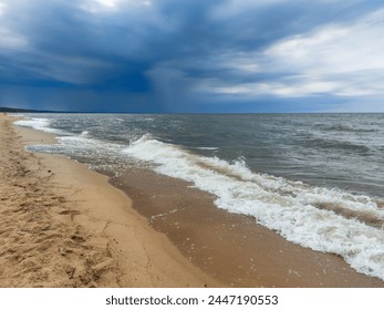 Seascape of the Baltic sea with big foamy waves in a windy day with yellow sand on the shore and very dark blue, contrasting clouds above Stock Photo
