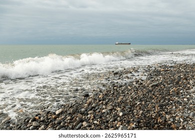 Seascape with blue and red oil tanker or cargo vessel on distant horizon, crashing wave splashing against pebble beach shore in foreground