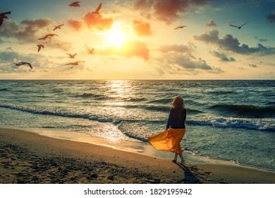 Seascape at sunrise with beautiful sky. Woman on the beach. Young happy woman in a yellow fluttering dress walks along the seashore. The girl looks at the magical sunrise. Seagulls fly over the beach