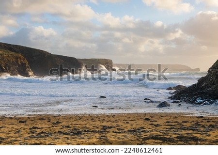 Seashore scene showing frothy white sea water rolling onto a Beach and waves crashing against rocks at Dollar Cove, The Lizard, Cornwall with a cloudy sky background