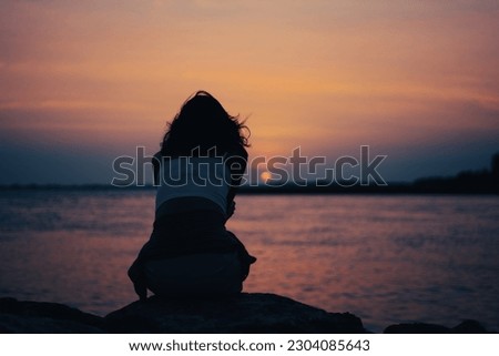 At the seaside in the evening, where the wind is so strong that her hair is blowing, a young woman is in the mood to watch the sun set