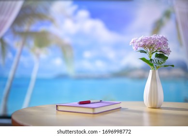 Seaside house interior wooden desk with notebook, pen, and a vase of purple flowers. Beautiful blurry view of beach landscape with blue sky and white clouds. Copy space.
