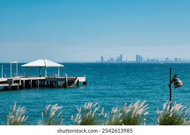 Seaside landscape with a wooden pier and white pavilion overlooking a turquoise sea, with a city skyline visible on the horizon. Tall grass waves gently in the foreground under a clear blue sky, creat - Powered by Shutterstock