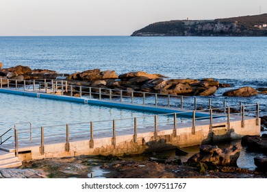 Seaside Ocean Swimming Pool With Metal Hand Rail Fencing Set Against Calm Blue Water And Large Exposed Rocks