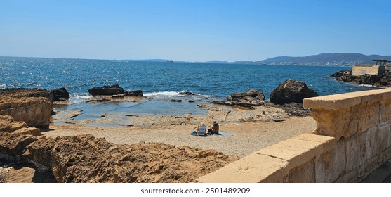 Seaside scene with two people sitting on a small rocky beach, calm blue sea, and distant mountains on the horizon. - Powered by Shutterstock