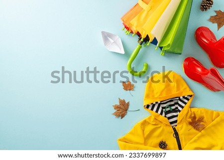 Seasonal bliss. Top view scene with child's yellow raincoat, red gumshoes, cheerful rainbow umbrella, paper boat, maple leaves, pine cones on pastel blue backdrop, leaving space for text or advert