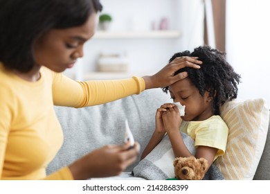 Seasonal Flu Concept. Worried Black Mom Taking Care Of Her Ill Child At Home, Caring Mother Touching Daughter's Forehead And Looking At Thermometer, Checking Kid's Temperature, Closeup Shot - Shutterstock ID 2142868399
