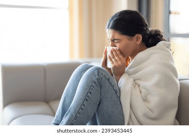 Seasonal Flu. Sick Young Indian Woman Blowing Nose In Paper Tissue While Sitting On Couch At Home, Ill Eastern Female Covered In Blanket Suffering Runny Nose, Got Cold, Side View With Copy Space