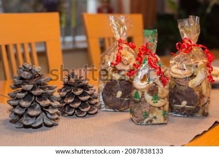 Seasonal homemade advent or christmas cookies in transparent bags gift wrapped with curled red ribbon placed on a table which is decorated with fir cones, bokeh lights, blurred background