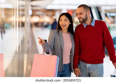 Seasonal Sales. Cheerful Interracial Couple Shopping For Clothes At Mall Centre, Looking At Store Window, Copy Space. Millennial Diverse Family With Shopper Bags Making Purchases At Supermarket