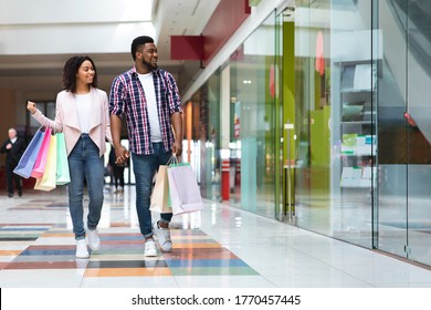 Seasonal Sales. Happy Black Couple Walking In Mall Center After Buying Clothes, Looking At Store Window, Copy Space
