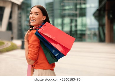 Seasonal Sales. Happy Stylish Young Woman Holding Lots Of Shopping Bags On Shoulder, Smiling To Camera Walking At Modern Mall Urban Area Outdoor, Standing Back To Copy Space