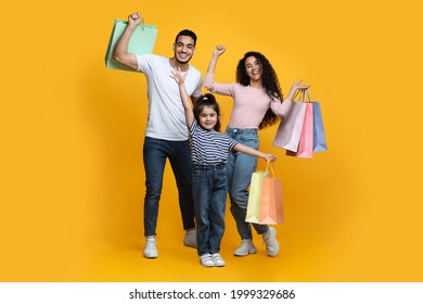 Seasonal Sales. Portrait Of Excited Happy Arab Family Of Three Carrying Shopping Bags And Raising Hands, Cheerful Young Middle Eastern Parents And Their Little Daughter Posing Over Yellow Background