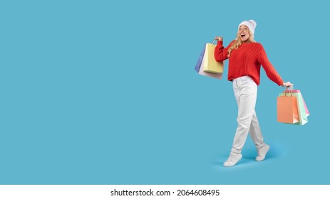Seasonal Sales And Shopping. Full length of overjoyed female consumer walking and looking at free copy space, lady holding many colorful bags with new purchases isolated on blue studio wall, advert